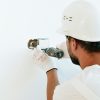 Construction worker wearing helmet and gloves uses a power tool to fix a wall.
